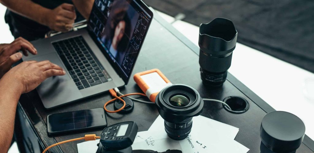 Photographer looking at photos after a photo shoot on a laptop. Photographer editing photos on laptop with camera and lens placed on table.